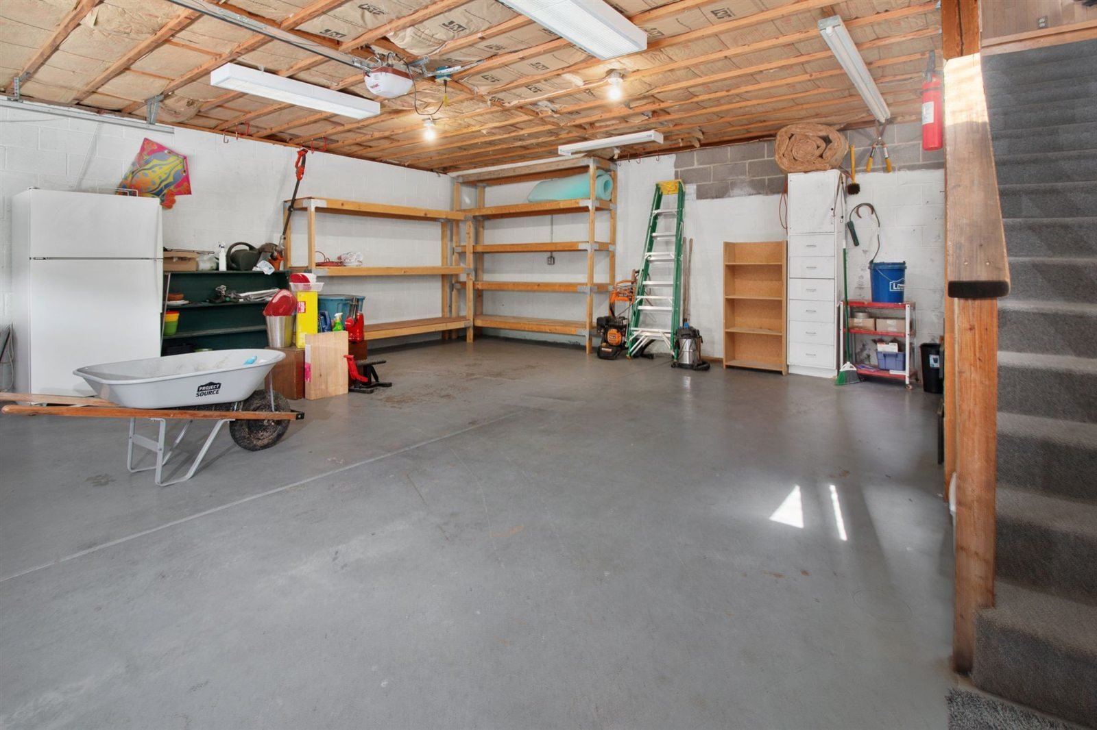 A residential garage interior with a bare grey concrete floor, shelving and a wheelbarrow